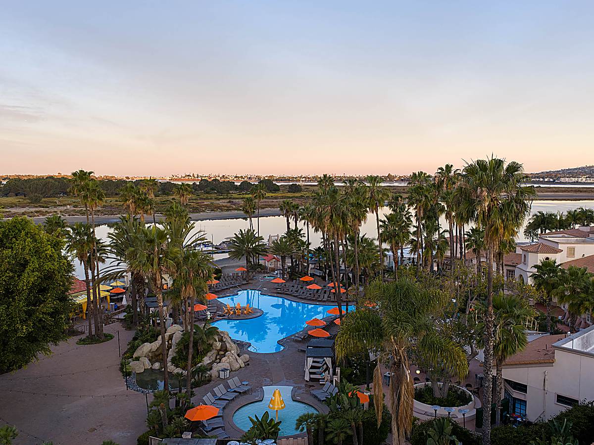 Sunset view of the San Diego Mission Bay Resort pool surrounded by palm trees, orange umbrellas, and lounge chairs overlooking Mission Bay