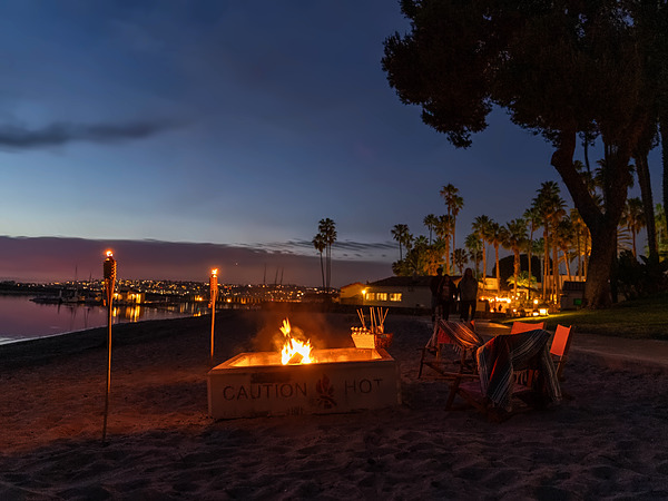 Beachfront fire pit at dusk with tiki torches and chairs on the sand at San Diego Mission Bay Resort.